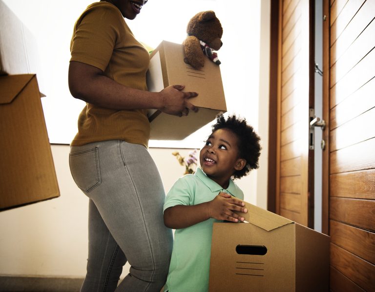 A young boy smiles as he holds a cardboard moving box as he walks toward a door.